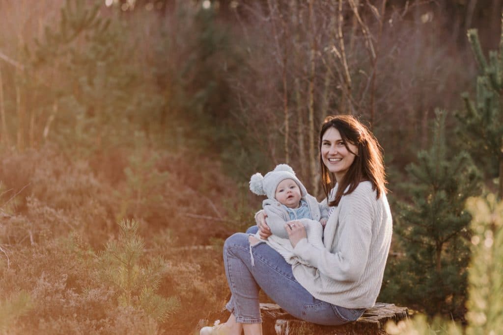 Mum is sitting on the tree stamp and looking at the camera. She is smiling and holding her baby boy. Family photographer in Hampshire. Ewa Jones Photography
