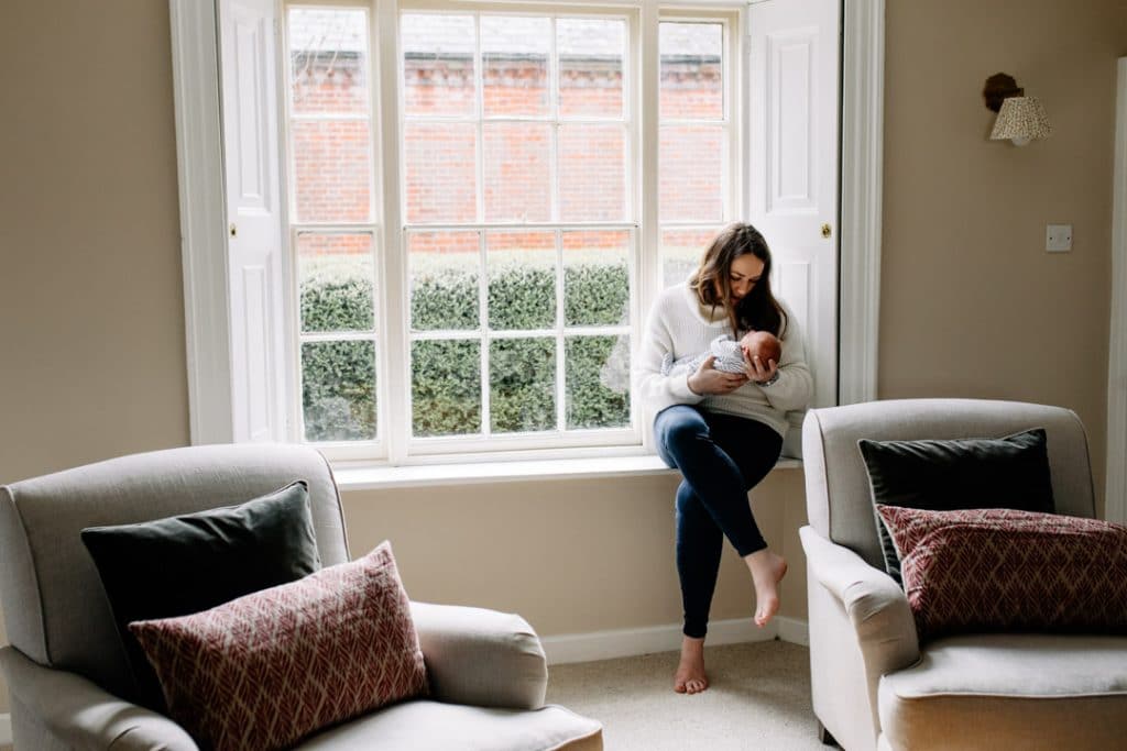 Mum is sitting on the window ledge and holding her newborn baby in her arms. Newborn photographer in Hampshire. Ewa Jones Photography