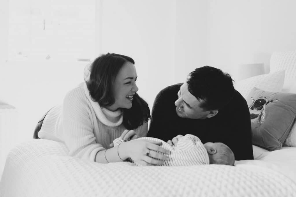 Mum and dad are laying on the bed and looking at each other. their newborn baby boy is laying on bed too. Newborn photoshoot in Hampshire. Ewa Jones Photography