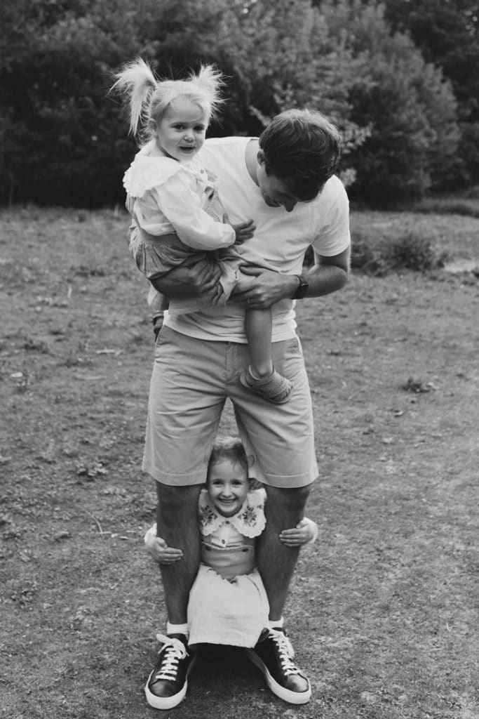 Black and white image of dad holding his daughter and another daughter is hiding behind his legs. Lifestyle family photography in Hampshire. Hampshire photographer, Ewa Jones Photography