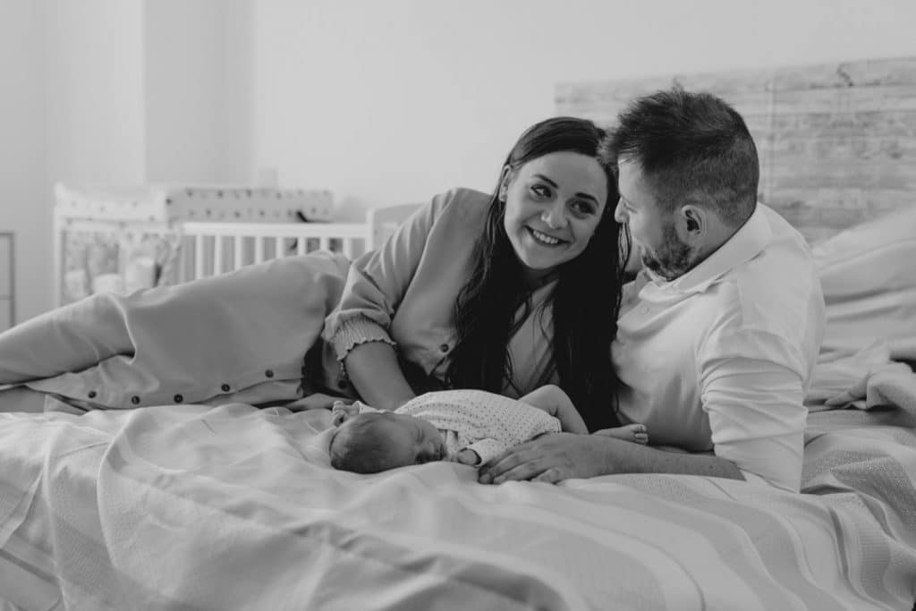 Mum and dad are laying on the bed and looking at each other. Their baby boy is sleeping on the bed. Hampshire photographer. Ewa Jones Photography