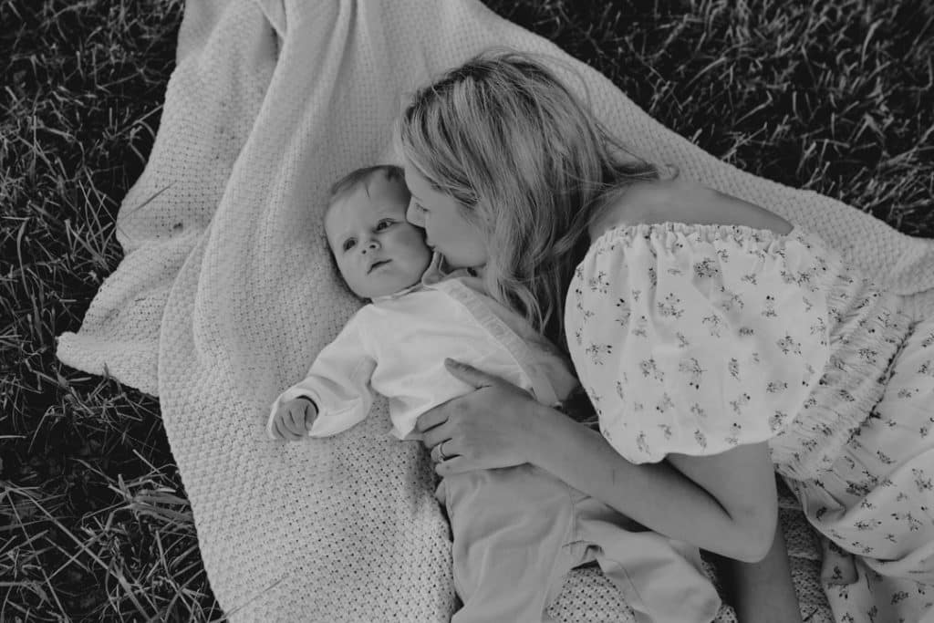 Family photo session in Hampshire countryside-11 Black and white image of mum and son laying on the blanket. Mum is kissing her baby boy. Natural lifestyle family photo session in Hampshire. Hampshire family photographer. Ewa Jones Photography