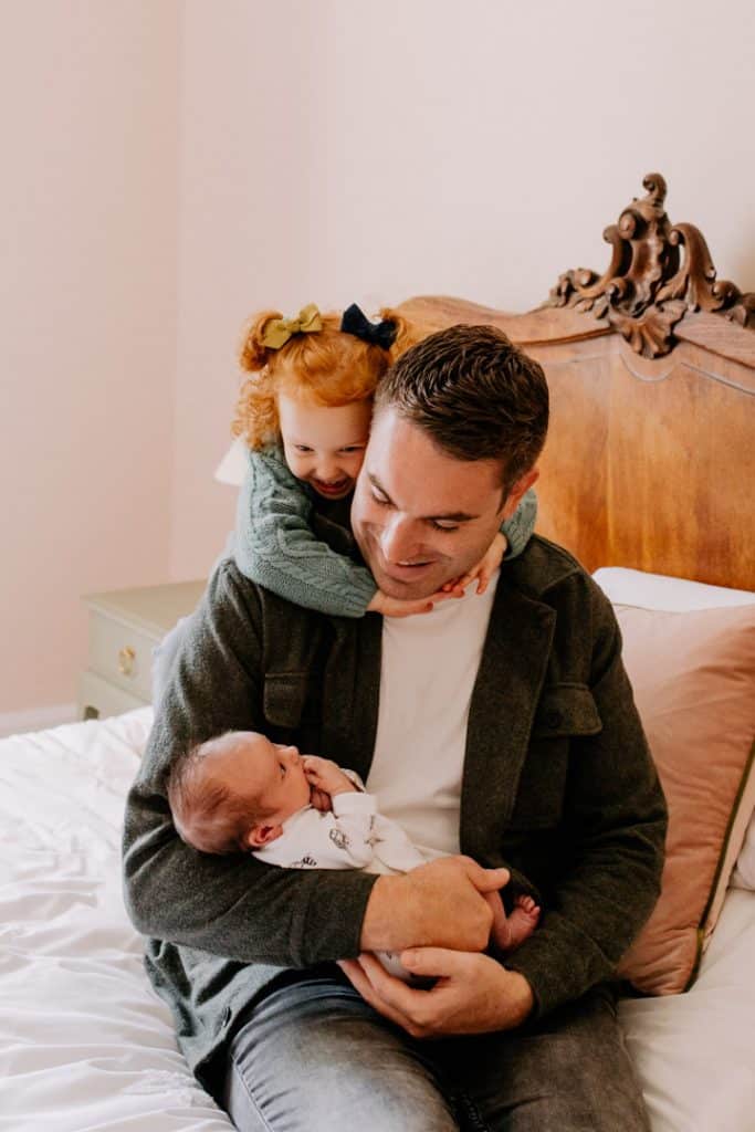 dad is sitting on the bed and holding his newborn baby girl. his older daughter is cuddling him. Newborn photo shoot in Berkshire. Newborn photographer. Ewa Jones Photography
