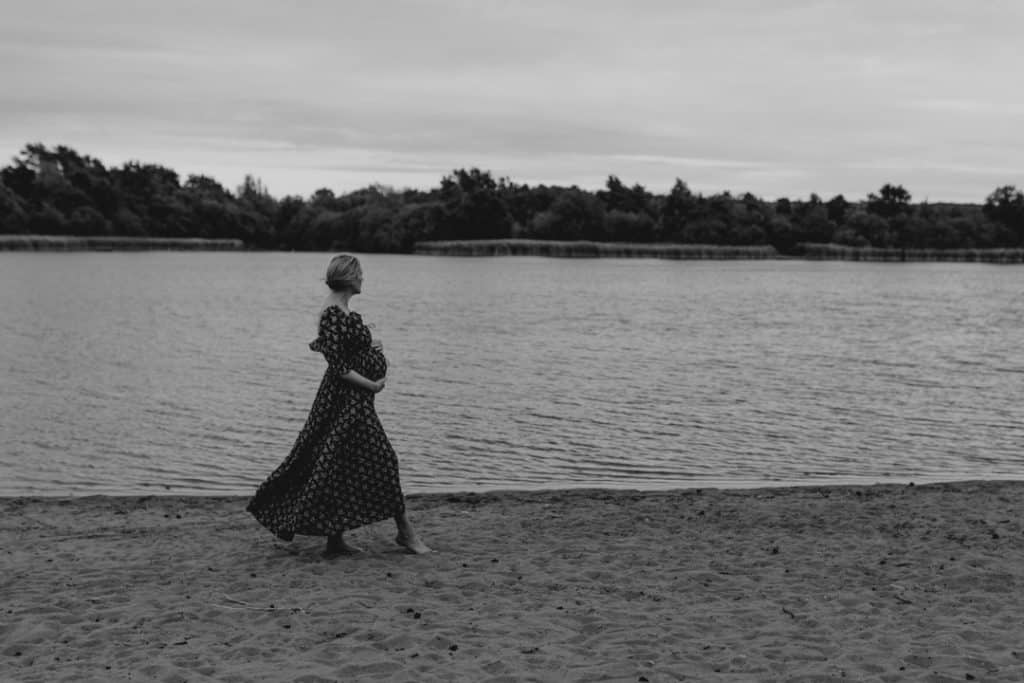 Pregnant mum is holding her bump. She is wearing lovely flower dress. She is standing by beautiful lake. Maternity photo session in Hampshire. Ewa Jones Photography