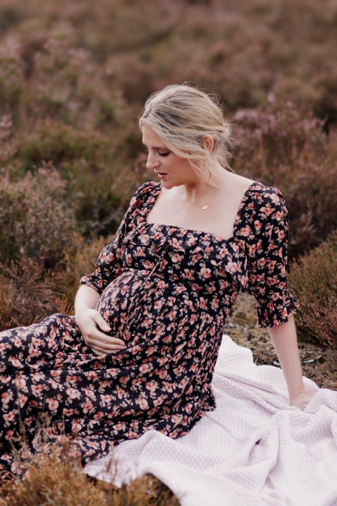 Pregnant mum is sittin gon the blanket and looking down at her baby bump. She is wearing lovely flowery dress. maternity poses. maternity photo session in Hampshire. Ewa Jones Photography