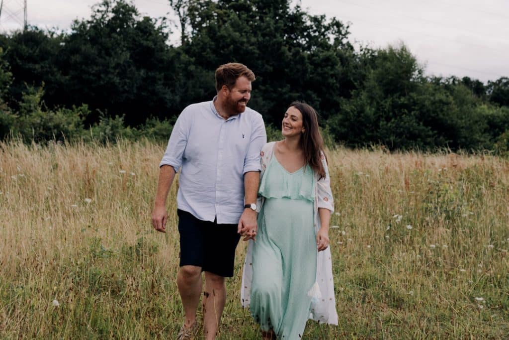 Pregnant mum and her husband are walking through the wild field and smiling. Lovely natural maternity photo session in Basigstoke. Ewa Jones Photography