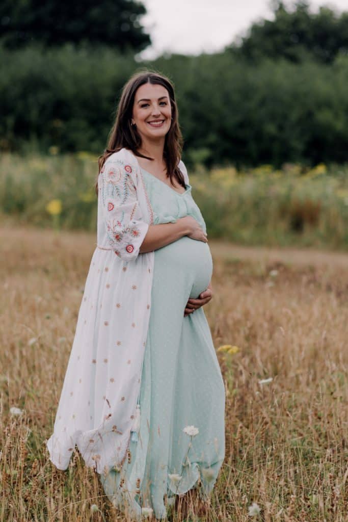 Pregnant mum is standing in the field and holding baby bump. She is wearing lovely summer dress. Matenrity photo shoot outdoor. Hampshire maternity photographer. Ewa Jones Photography