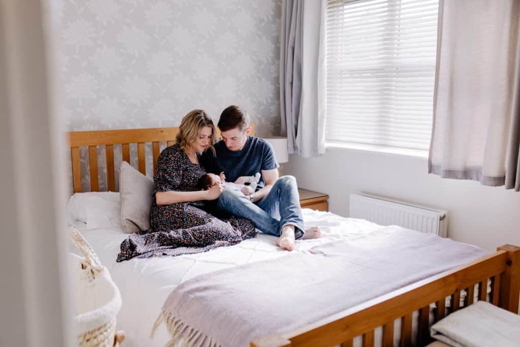 mum and dad are sitting on the bed and cuddling their newborn baby girl. Newborn photo shoot at home. Hampshire newborn photographer. Ewa Jones Photography