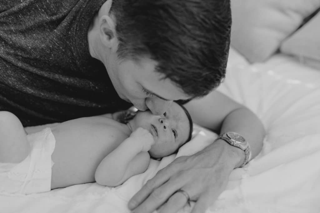 Dad is kissing his newborn baby girl and the baby girl is laying on bed. Newborn photo session in Basingstoke Hampshire. Ewa Jones Photography