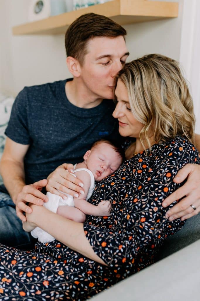 Mum and dad are cuddling close and dad is kissing mum. Newborn baby is laying on mums chest. Newborn photo session in home. newborn photographer in Hamsphire. Ewa Jones Photography