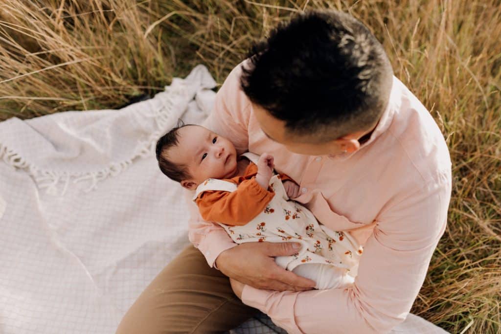 Dad is holding his baby girl and looking at her. Baby girl is looking at dad. Family photo session outdoors. Lifestyle family photo session. Ewa Jones Photography