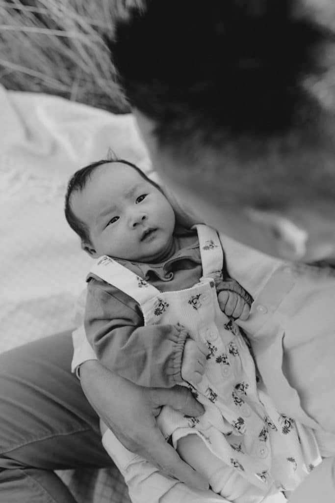 Dad is holding his baby girl and looking at her. Baby girl is looking at dad. Family photo session outdoors. Lifestyle family photo session. Ewa Jones Photography