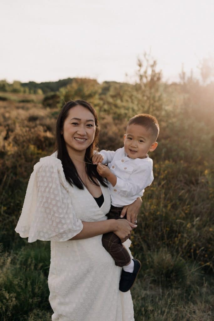 Mum is looking at camera and smiling. her little boy is looking at the camera and smiling too. Lovely golden hour family photo shoot. Family photo session outdoors. Lifestyle family photo session. Ewa Jones Photography