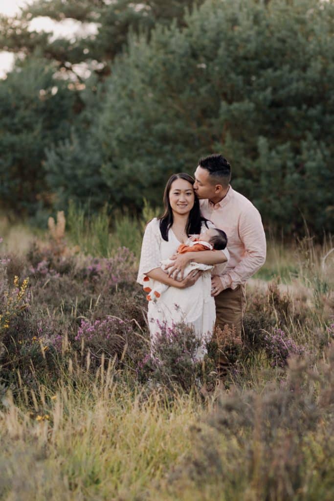 Mum and dad are standing in the field and dad is holding mum and kissing. Mum is holding her baby girl in her arms who is sleeping. family photo session in Hampshire. Ewa Jones Photography