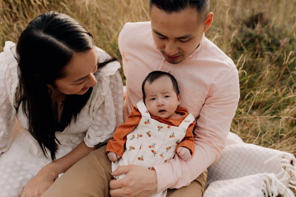 family is sitting on the blanket and mum is looking at her baby girl. Dad is also looking down at baby girl. Family photo session in Surrey. Ewa Jones Photography