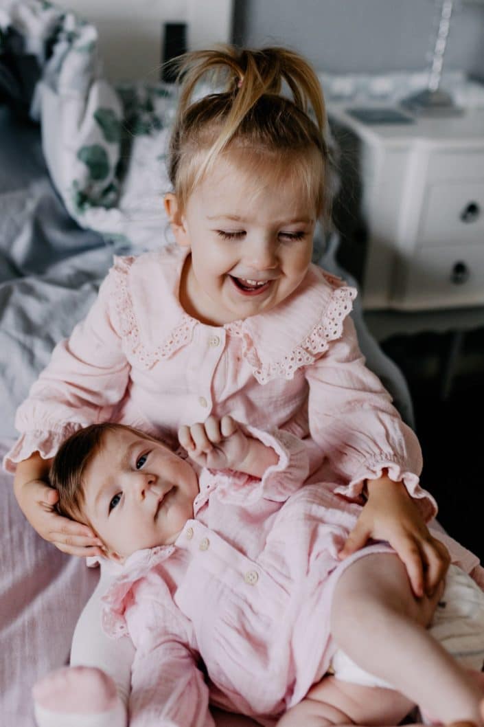 Newborn baby girl is laying and looking at the camera and her sister is cuddling her. Lovely image of two sisters. Newborn baby photo shoot in Hampshire. Newborn photography Hampshire. Ewa Jones Photography