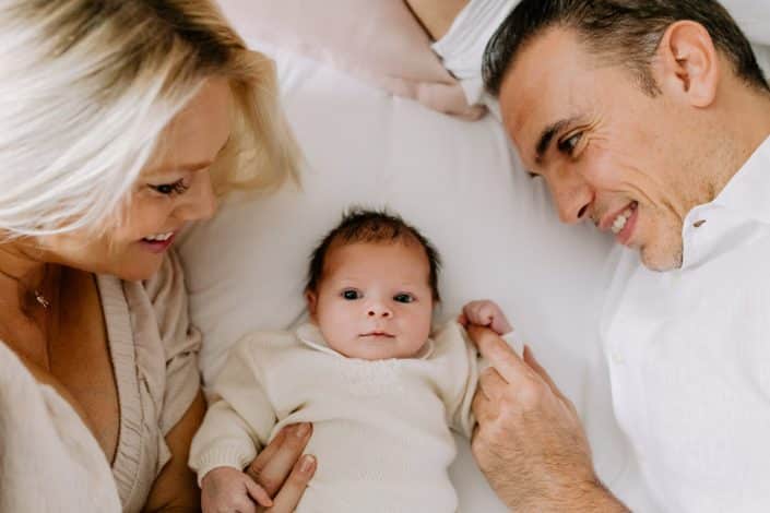 Mum and dad are laying on the bed and looking at their baby girl. She is smiling and looking at the camera. Newborn photography in London. Hampshire photographer. Ewa Jones Photography