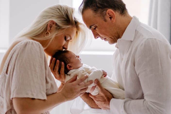 Mum is kissing her baby girl on her head and dad is looking down and her baby girl. Lifestyle newborn photo shoot in London. Hampshire newborn photographer. Ewa Jones Photography