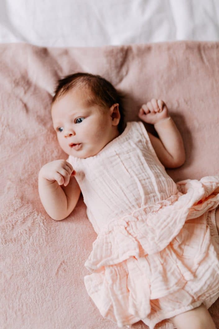 Newborn girl is laying on the bed and looking away. She is wearing lovely pink dress. Newborn photo shoot in London. Hampshire photographer. Ewa Jones Photography