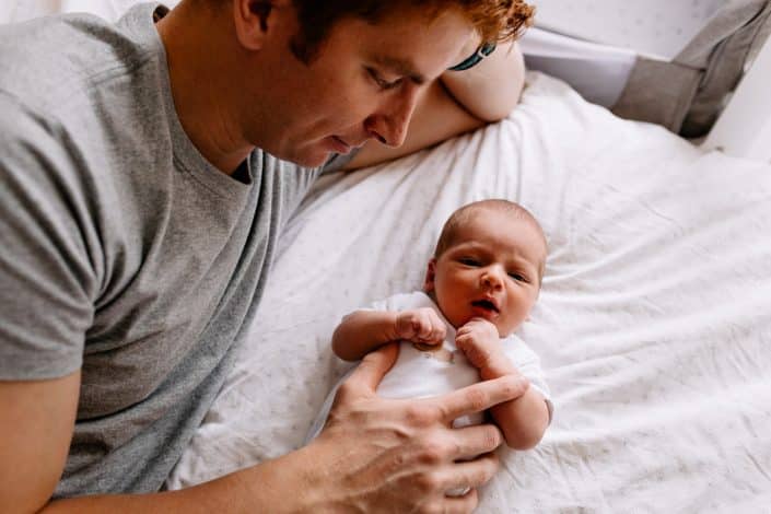 Newborn baby boy is laying on the bed and looking at the camera. Dad is laying next to him. Newborn photoshoot in Hampshire. Hampshire newborn photographer. Ewa Jones Photography