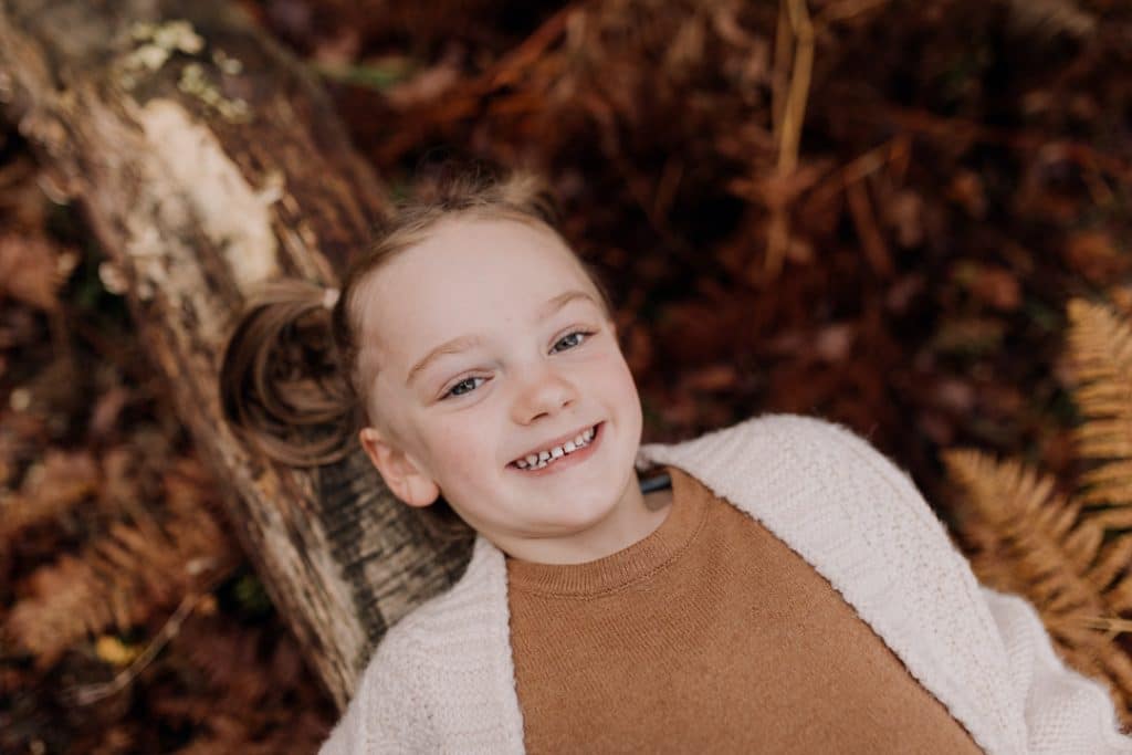 Young girl is laying on the log and looking at the camera. she is wearing lovely brown dress and a cardigan. Family photographer in Hampshire. Hampshire family photo session. Ewa Jones Photography