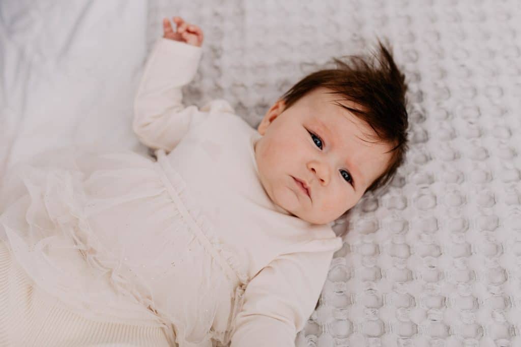 baby girl is laying on the bed. she is wearing lovely cream dress and looking at the camera. natural photo session at home. hampshire photographer. Ewa Jones Photography.
