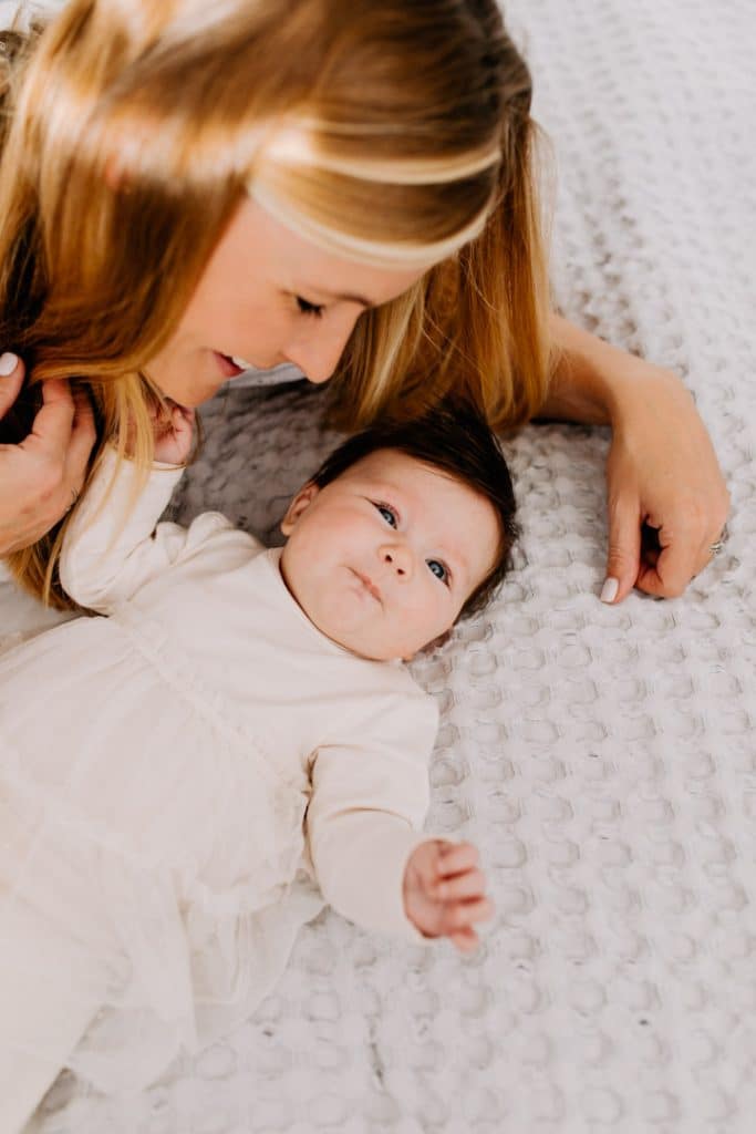 mum is laying on the bed and looking at her baby daughter. baby is looking at the camera. she is wearing lovely cream dress. Newborn photo shoot at home. Ewa Jones Photography.