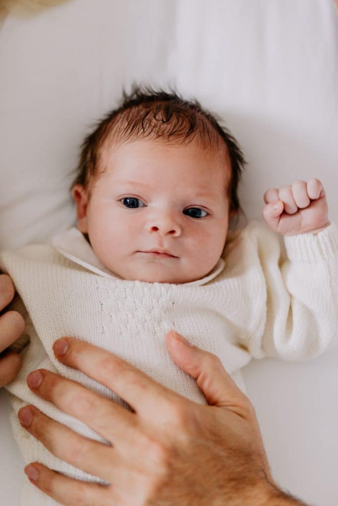 Newborn photo shoot in London-5 Newborn baby girl is laying on the bed and dad has his hands on her tummy. She is wearing lovely cream jumper. Natural and lifestyle photo session in London. Newborn photographer in Basingstoke. Ewa Jones Photography