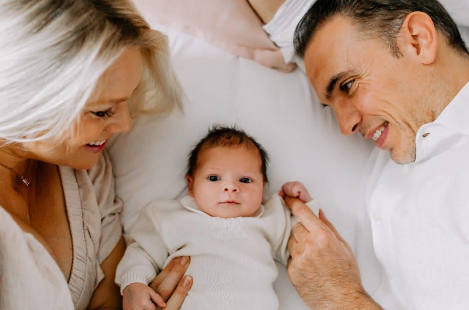 Mum and dad are laying on the bed and looking at their baby girl. Baby girl is looking at the camera. newborn photo session. Hampshire newborn photographer. Ewa Jones Photography. Newborn photoshoot in London