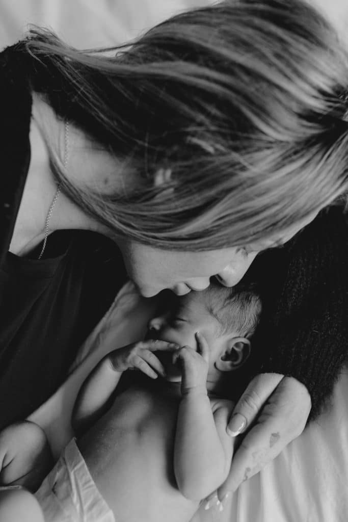 Mummy is kissing gently her newborn baby girl on her head. baby girl is sleeping. Hampshire newborn photo session at home. newborn baby girl is laying on bed sleeping. Hampshire newborn photographer. Ewa Jones Photography