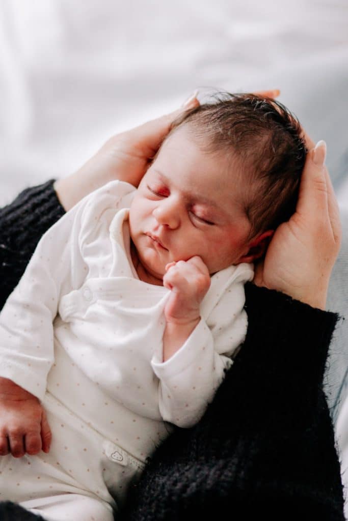 Newborn baby girl is being held by her mum. she is wearing lovely white outfit. Newborn photo shoot at the comfort of your own home. Hampshire newborn photo session. Ewa Jones Photographer
