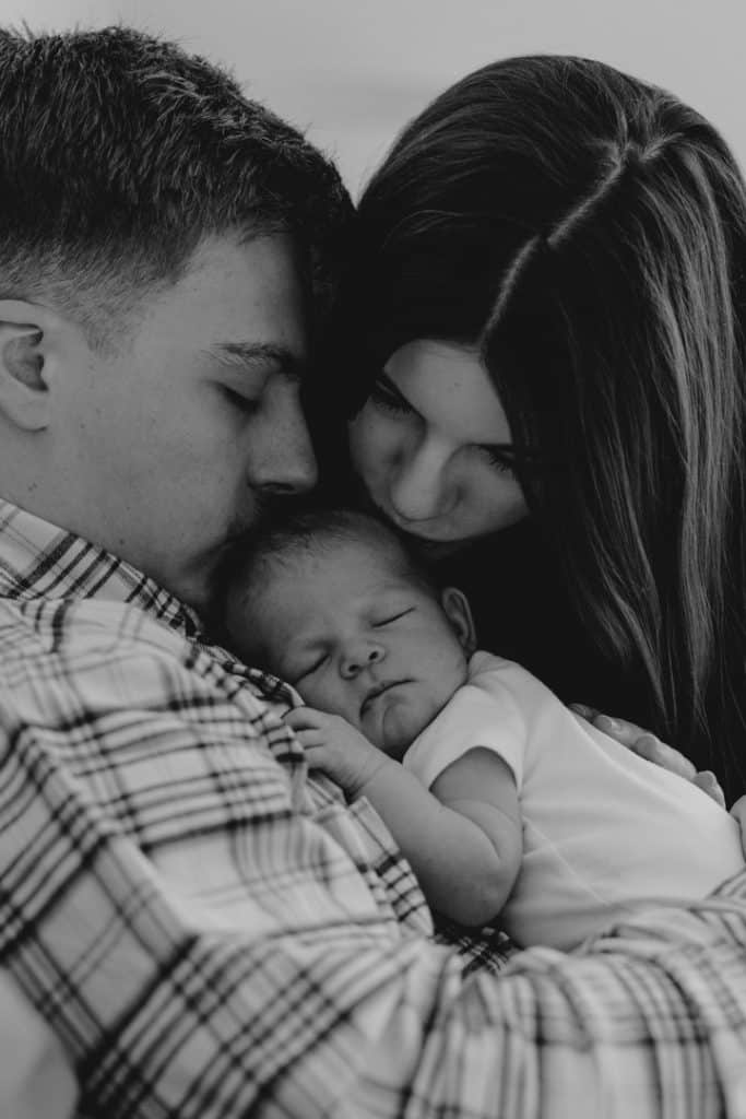 mum and dad are kissing their newborn baby girl. newborn baby is sleeping on daddy. candid newborn photo shoot at home. Newborn photographer in Hampshire. Ewa Jones Photography