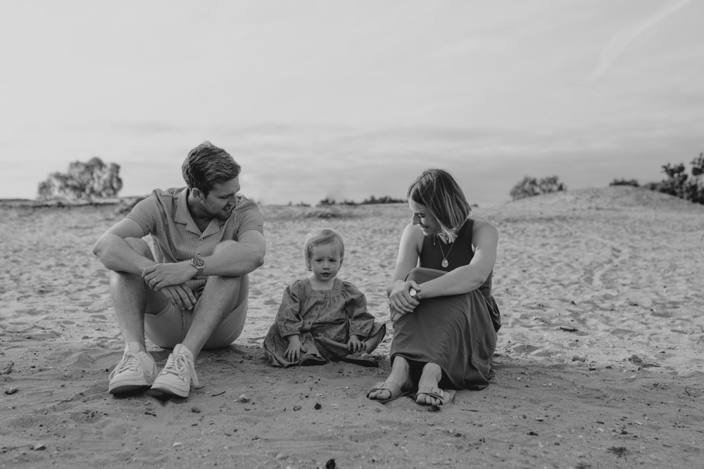 family of three is sitting on the sand and mum and dad are looking at their daughter. family photo session is six steps. Ewa Jones Photography