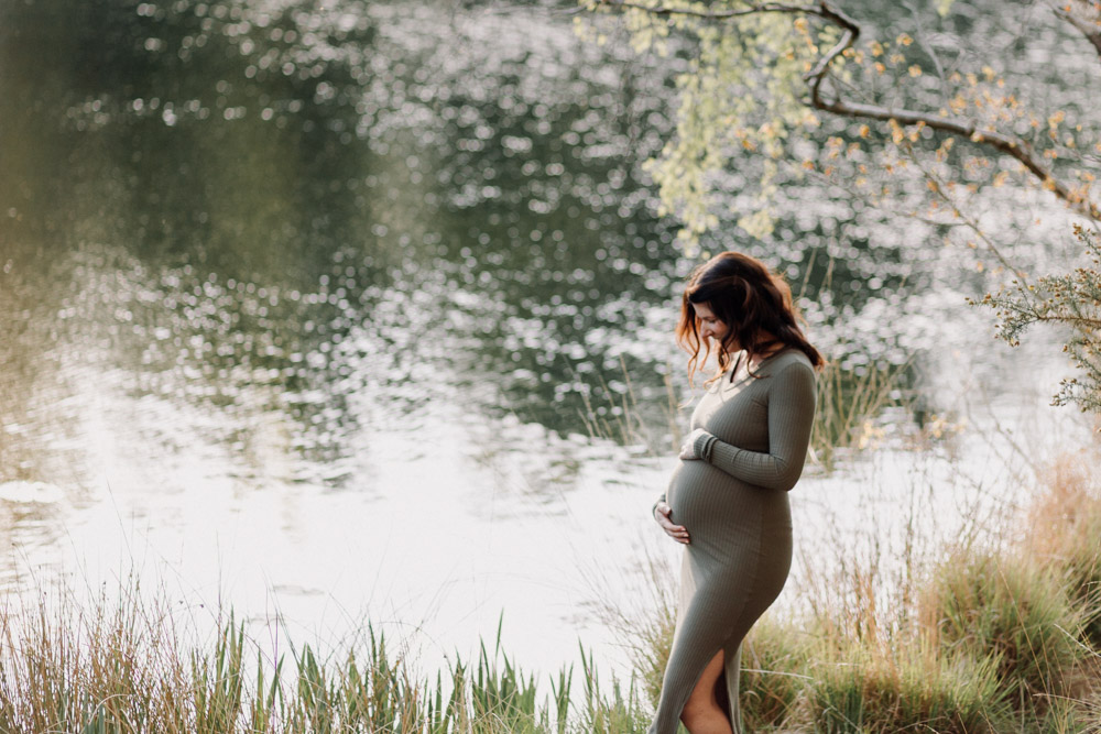 Mum is looking down at her bump. Lovely scenery. Nature reserve. Pond in the background. Ewa Jones Photography