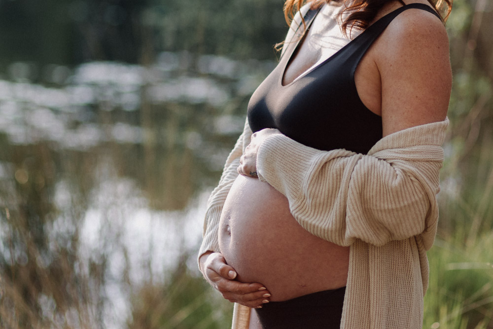 Mum is looking down at her bump. Lovely scenery. Nature reserve. Pond in the background. Ewa Jones Photography