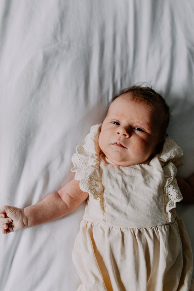 Baby girl is looking at the window. She is wearing lovely cream baby grow. Newborn photo shoot at home. Basingstoke photographer. Ewa Jones Photography