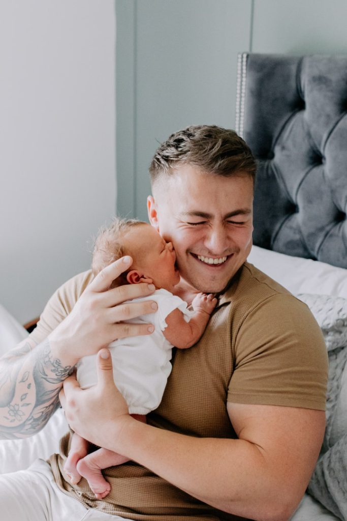 newborn baby girl is trying to kiss her dad. Dad is sitting on the bed and laughing. Newborn photo session at home. Hampshire photographer. Ewa Jones Photography