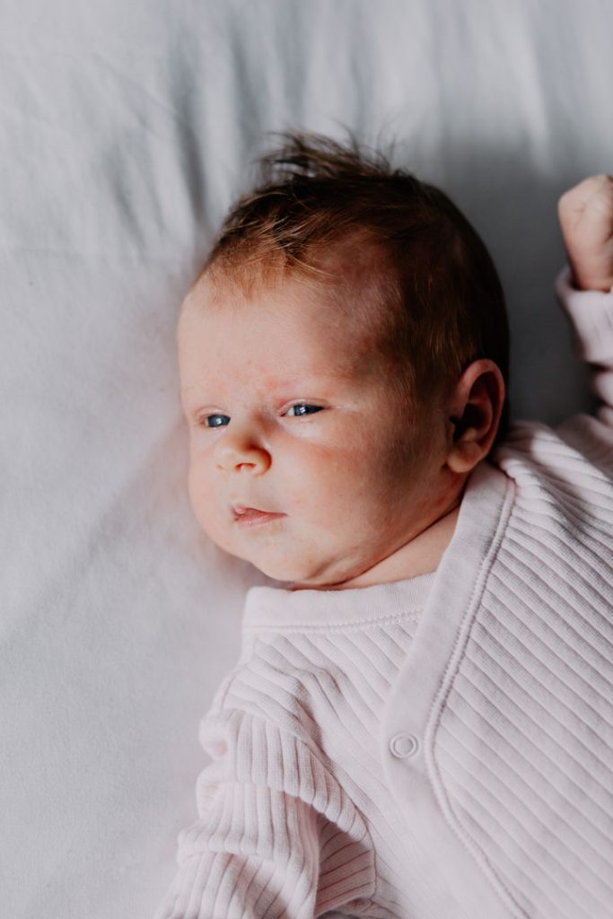 Baby girl is looking at the window. She is wearing lovely light pink baby grow. Newborn photo shoot at home. Basingstoke photographer. Ewa Jones Photography