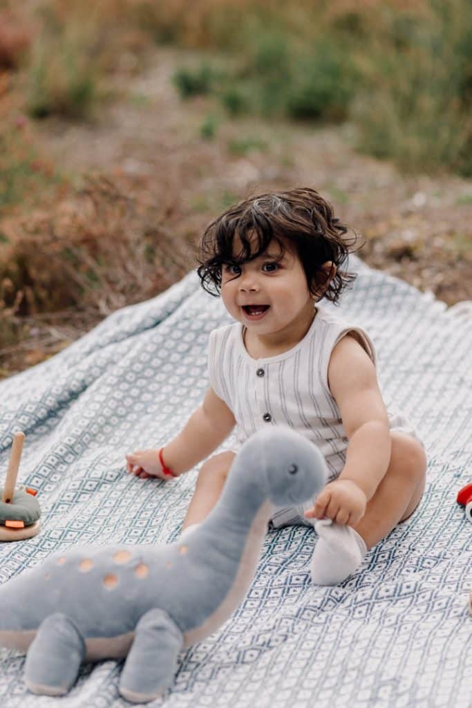 Little boy is sitting on the blanket. He is smiling and has his toys around him. Celebrating one year family photo session. Family photographer. Ewa Jones Photography