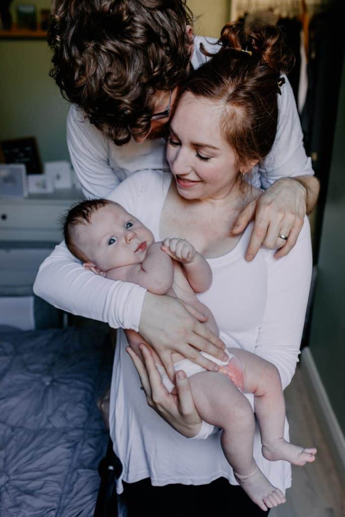 Mum and dad are cuddling together and looking down at the baby boy. baby boy is looking at the camera. In home intimate newborn photo shoot. Hampshire photographer. Ewa Jones Photography