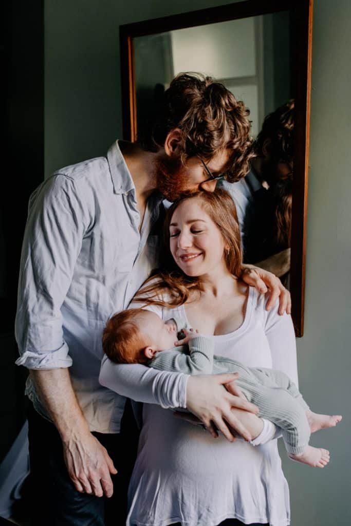 Mum and dad are standing and dad is kissing mum on top of her head. Mum is holding their baby boy. Newborn photography. Newborn photographer in Hampshire. Ewa Jones Photography