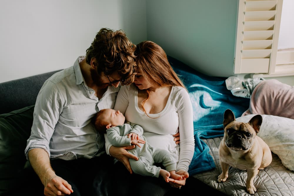 mum and dad are sitting on the sofa and looking down at their baby boy. Newborn photo shoot at home. Ewa Jones Photography