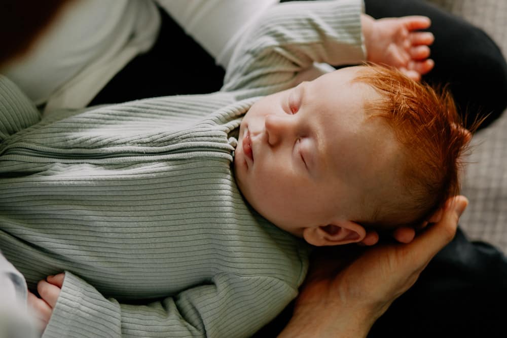 Baby boy is sleeping and dad is holding him in his arms. Baby boy is wearing nice green baby grow. Newborn poses ideas at home. Hampshire photographer. Newborn photographer. Ewa Jones Photography