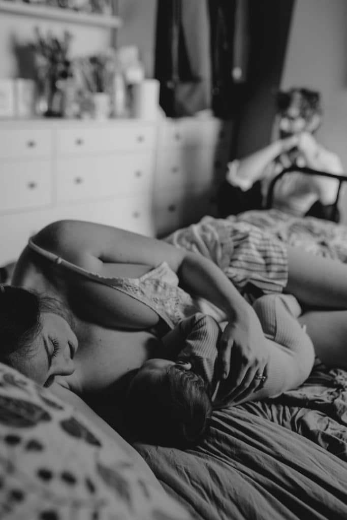 mum is breastfeeding her baby boy and laying on the bed with the boy. dad is in the background. newborn in home photoshoot. Hampshire newborn photographer. Ewa Jones Photography