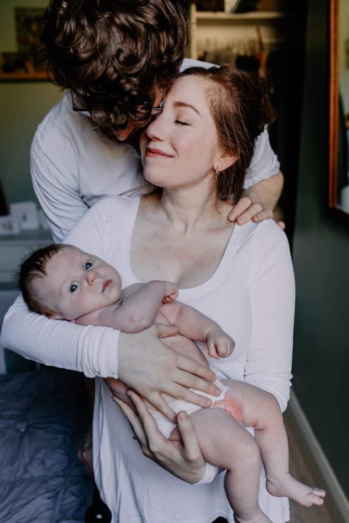 Mum and dad are cuddling together. Mum is holding a baby boy. Lovely in home natural newborn photo session. Hampshire photographer. Ewa Jones Photography