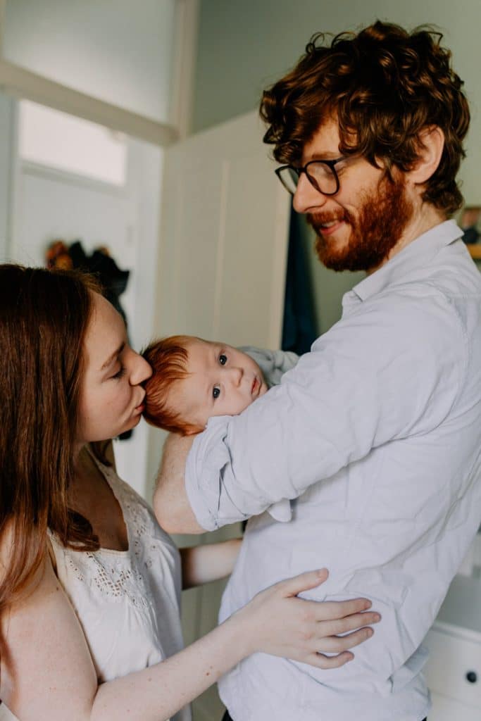 Mum and dad are standing and dad is holding their baby boy. Mum is kissing their baby boy on his head. Hampshire newborn photo session. Ewa Jones Photographer