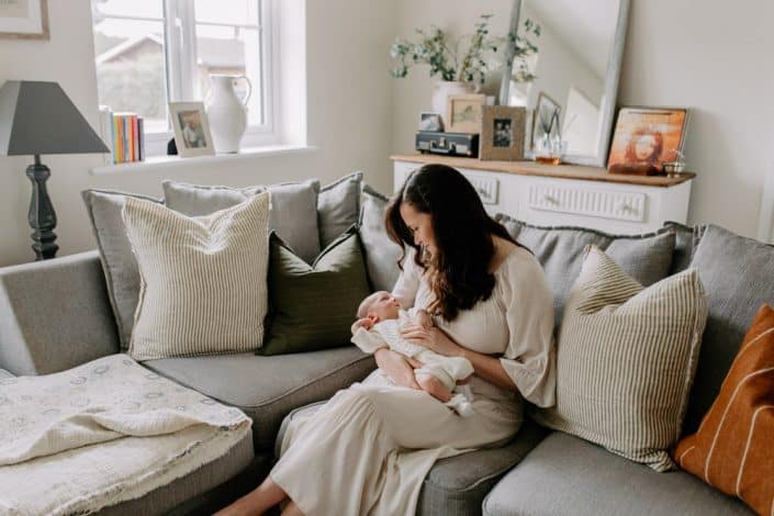 Mum is sitting on the sofa and holding her newborn baby in her arms. Baby boy is looking back at her. Newborn shoot at home. Natural baby photo shoot in your home. Hampshire photographer. Hampshire newborn photographer. Ewa Jones Photography