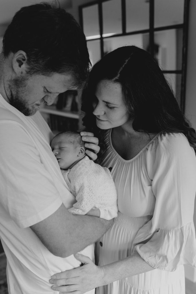 Lovely black and white image on dad holding his newborn baby. Mum is looking at the baby and cuddling in. Newborn photo shoot at home. Basingstoke photographer. Ewa Jones Photography