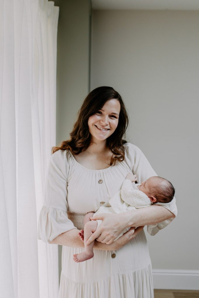 Mum is standing next to the window and smiling at the camera. she is holding her baby boy. lifestyle at home newborn photo shoot. Ewa Jones Photography