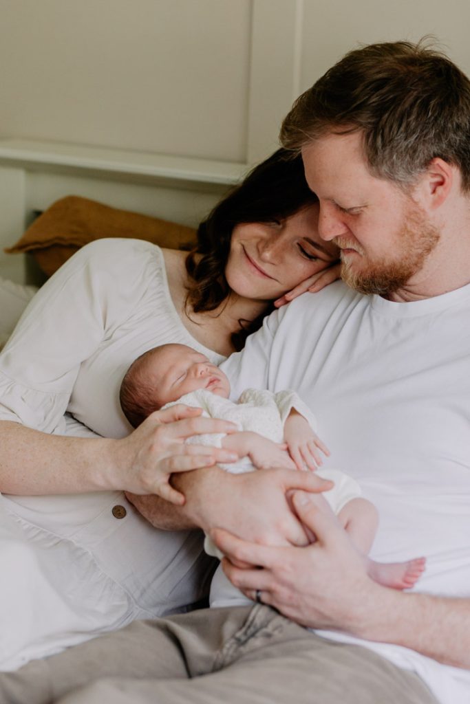 Mum is cuddling to her husband and looking down at her lovely newborn baby. Newborn baby poses during newborn photo session. Ewa Jones Photography